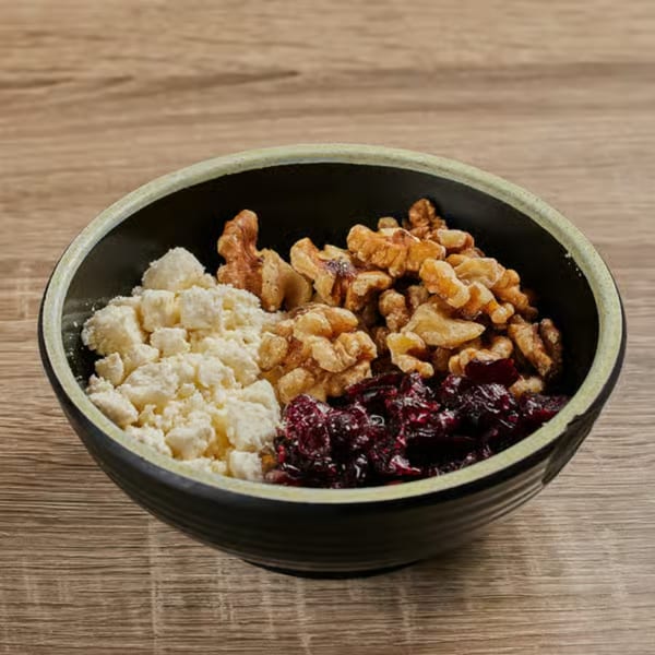 A black bowl filled with feta cheese, walnuts, and dried cranberries on a wooden table.