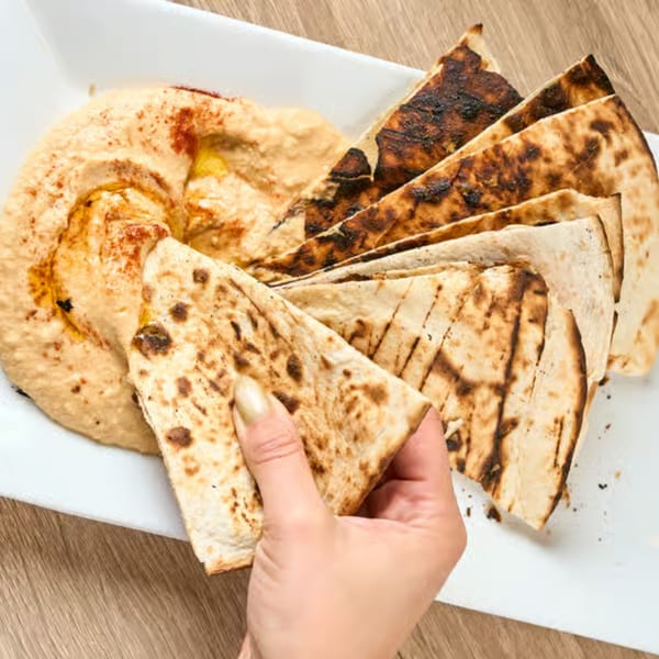 A hand dips a piece of grilled flatbread into a creamy bowl of hummus, garnished with spices.