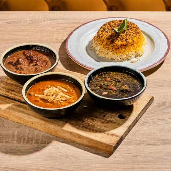 Three bowls of Persian stews and a plate of saffron rice with lime on a wooden board.
