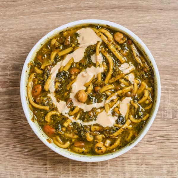 A bowl of Persian Ash Reshteh soup with noodles, herbs, beans, and a yogurt swirl.