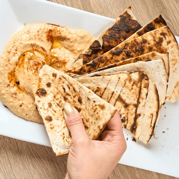 A hand dips a piece of grilled flatbread into a bowl of hummus on a white platter.