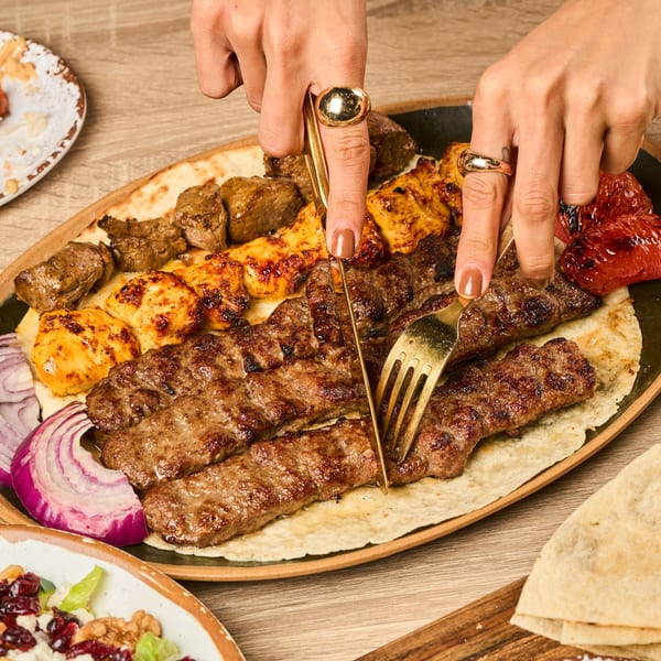 Hands cutting into a platter of Persian kabobs, including koobideh and chicken, with flatbread and onions.
