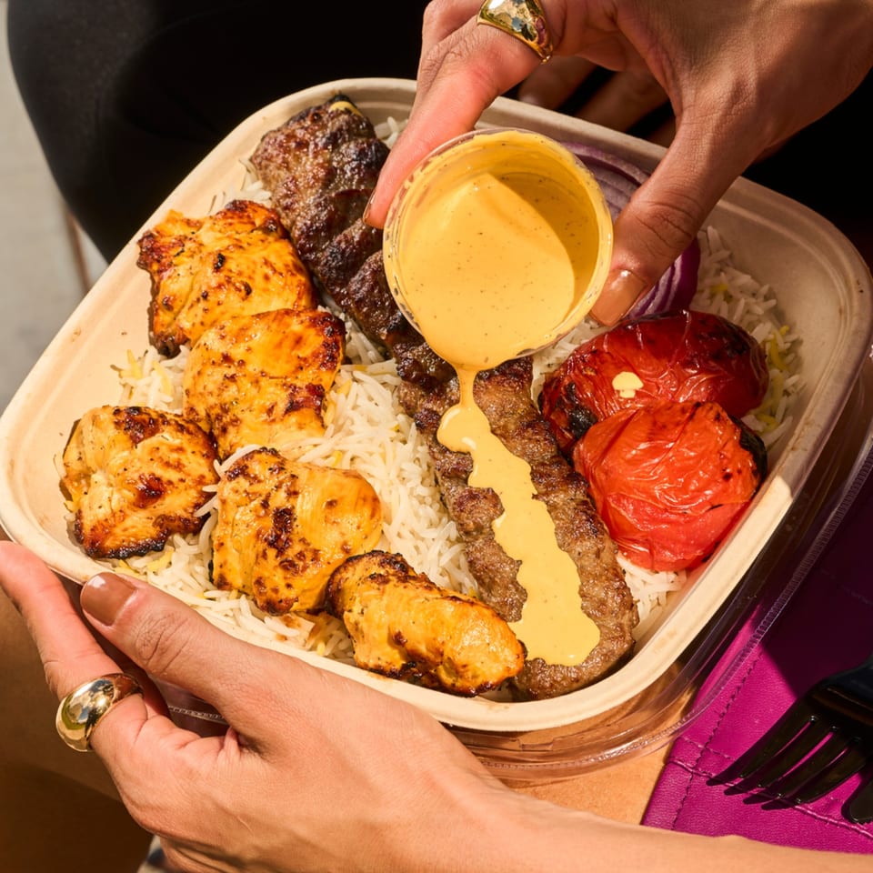 Hands holding a Shahs of Kabob takeout container with chicken, koobideh, rice, and grilled tomatoes.