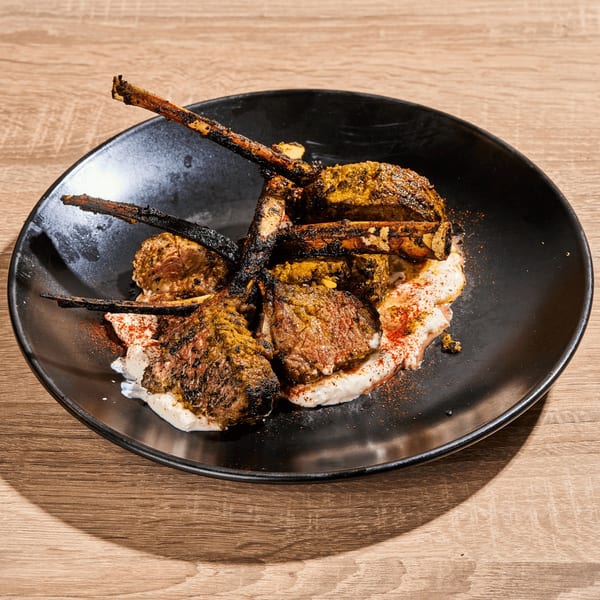 Grilled lamb chops with charred bones, spices, and a creamy white sauce on a black plate.